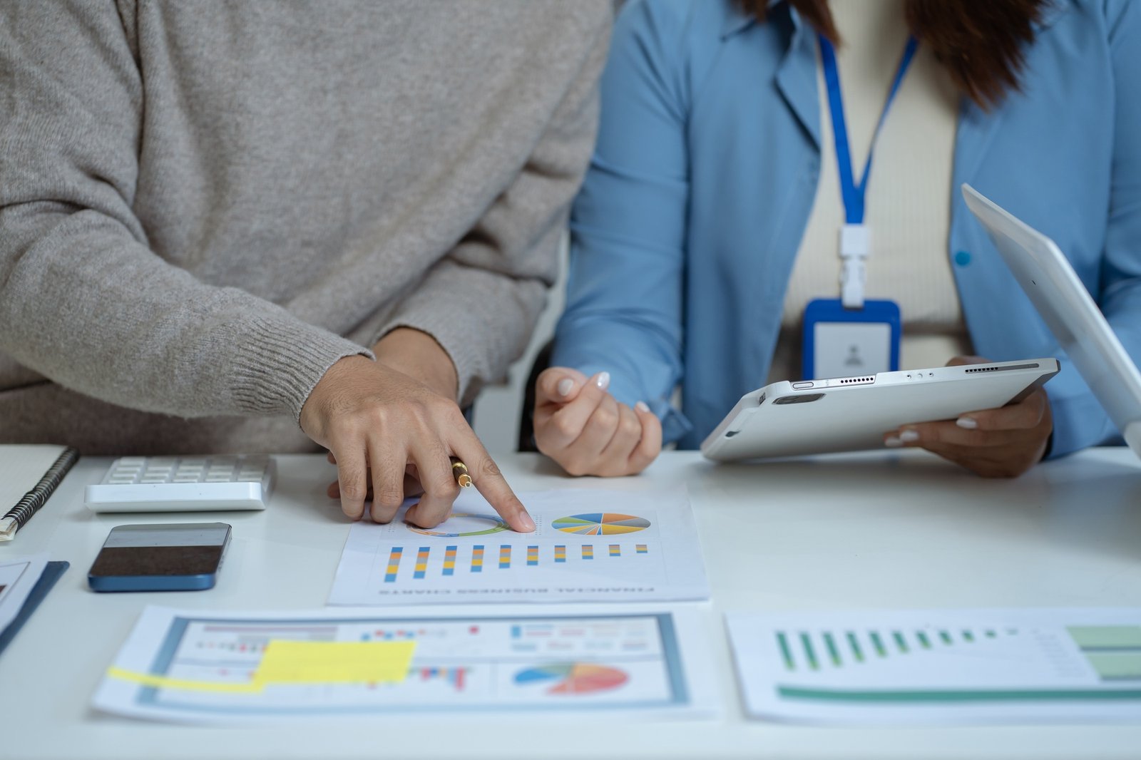 Dos personas señalando gráficos impresos sobre una mesa, una de ellas sostiene una tablet y ambas están parcialmente visibles.