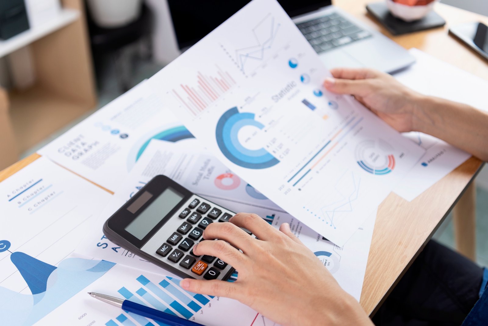 Persona usando una calculadora mientras observa gráficos impresos sobre una mesa, con diversos papeles y una laptop.