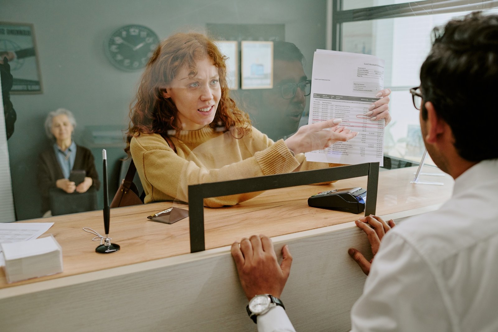 Mujer detrás de un mostrador señalando un documento que muestra a un hombre de pie frente a ella; se observa una división de vidrio.